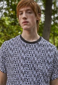 Camiseta gráfica en blanco y negro con logotipos de "Levi's" repetidos. El cuello redondo negro y las mangas cortas crean un diseño casual.