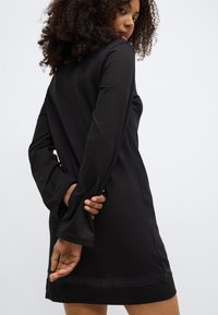 Črna obleka z dolgimi rokavi z ohlapnim krojem, z diskretno logotipno obrobo na robu in mehkim, gladkim materialom. Standardna dolžina.