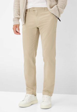 Beige Baumwoll-Chinos mit schmalem Schnitt, seitlichen Taschen und Knopfverschluss, kombiniert mit weißen Turnschuhen vor einem neutralen Hintergrund.