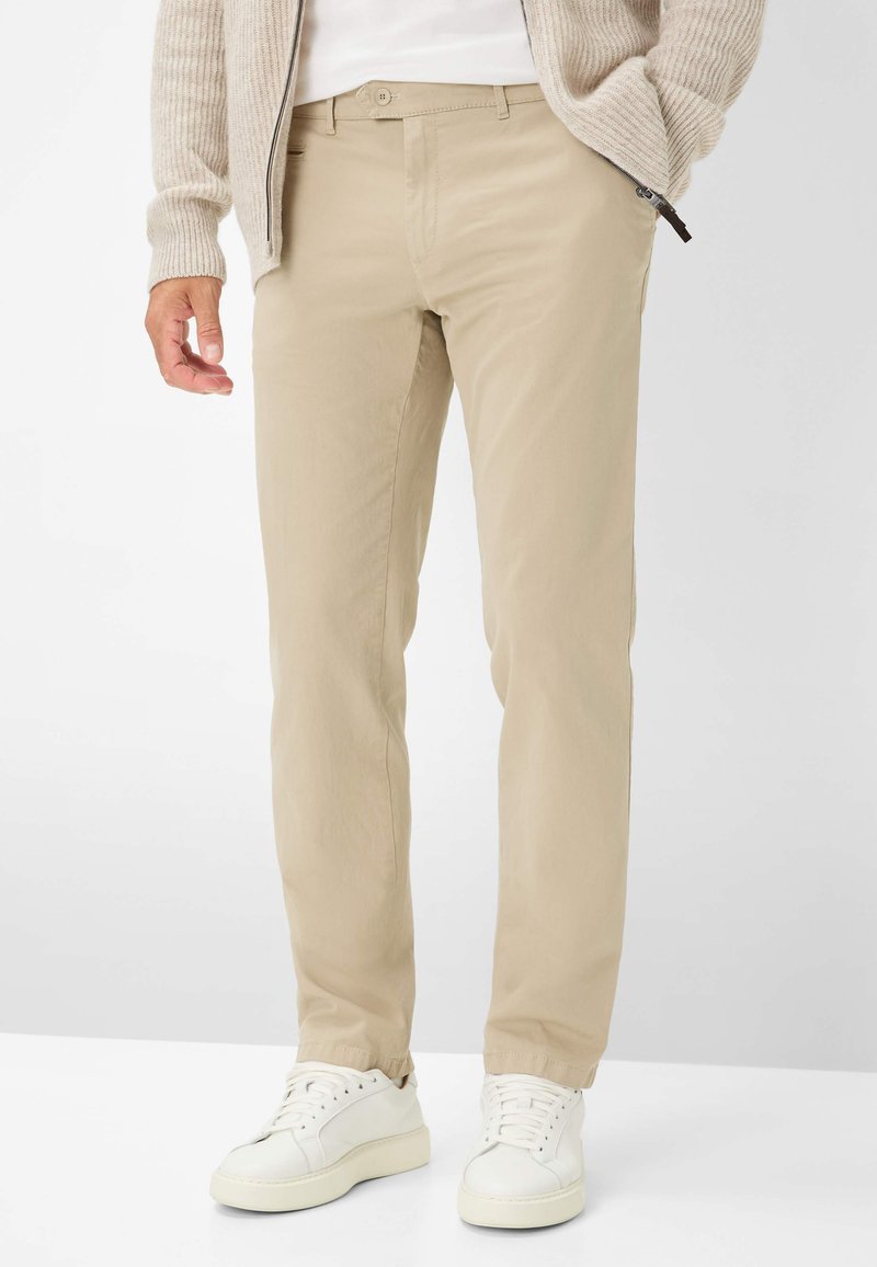 Beige katoenen chinos met een slim fit, steekzakken en een knoopsluiting, gecombineerd met witte sneakers op een neutrale achtergrond.