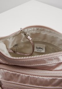 Otwarta torba w kolorze jasnoróżowym z satyny, z beżowym paskowanym wyściółką, kieszonką na zamek, klipsem na klucze oraz metką marki Kipling wewnątrz.