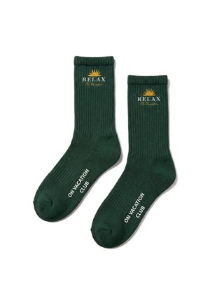 Chaussettes côtelées vert foncé avec les textes "RELAX On Vacation" et "ON VACATION CLUB" et un graphique de soleil jaune sur chaque chaussette.