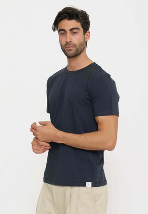 Uomo con capelli scuri che indossa una maglietta blu navy a maniche corte e pantaloni beige, in piedi contro uno sfondo chiaro e uniforme.