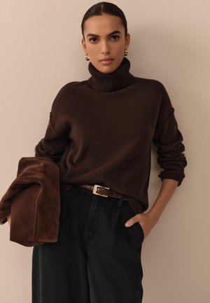 Brauner gestrickter Rollkragenpullover kombiniert mit hoch taillierten schwarzen Hosen und einer braunen Wildlederjacke, die einen strukturierten Gürtel und goldene Akzente aufweist.