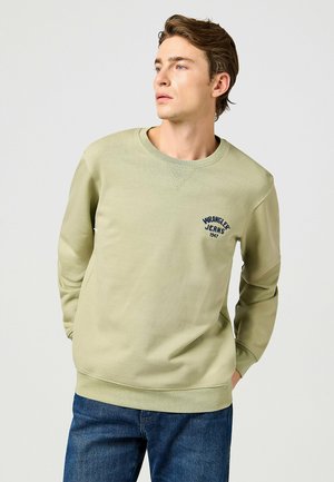 Felpa verde chiaro con collo rotondo, polsini e orlo a costine. Presenta un logo blu "Wrangler Jeans 1947" sul petto. Indossata con jeans blu.
