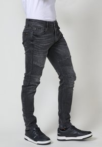 A fekete farmernadrág slim fit stílusú, megkopott textúrájú, és bordás részletekkel rendelkezik a comboknál, fekete magas szárú sneaker cipőkkel párosítva.