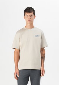 Beige Baumwoll-T-Shirt mit kurzen Ärmeln, Rundhalsausschnitt und blauer gedruckter Aufschrift "SOHO Social Coffee CLUB" auf der linken Brust. Glatte Textur.