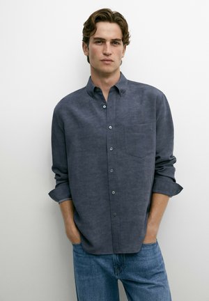 Giovane uomo con capelli castani corti e mossi indossa una camicia grigio scuro a maniche lunghe con bottoni e jeans blu, in piedi davanti a uno sfondo bianco.