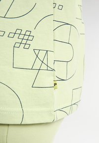 Lichtgroene t-shirt met blauwe geometrische lijnpatronen. Korte mouwen met een zichtbare stikselafwerking en een klein geel merklabel.