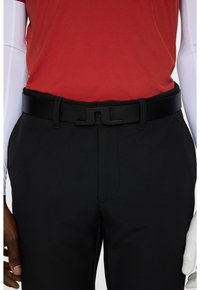 Pantaloni neri con una texture liscia, abbinati a una camicia rossa e una cintura nera con una chiusura distintiva e accenti di design eleganti.