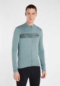 Maillot de cyclisme à manches longues en teal avec un tricot texturé, présentant une bande noire horizontale et un logo sur la poitrine, fermeture éclair.