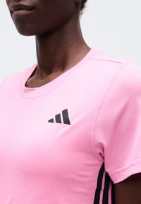 Γυναίκα που φοράει ροζ αθλητική μπλούζα με μαύρο λογότυπο τριών ρίγων στο στήθος και μαύρη ριγωτή λεπτομέρεια στο πλάι, μπροστά σε μονόχρωμο φόντο.