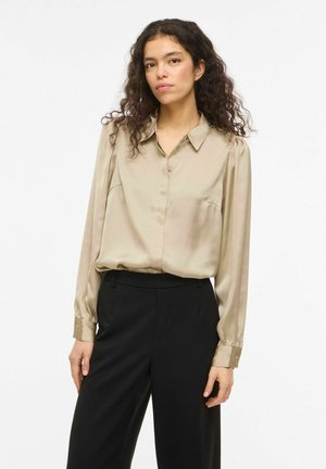 Kobieta o kręconych włosach, ubrana w beżową koszulę z długim rękawem, włożoną do czarnych spodni z wysokim stanem, stojąca na białym tle.