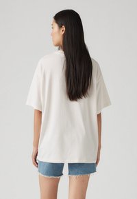 Camiseta blanca de gran tamaño con mangas cortas, hecha de algodón, combinada con pantalones cortos de denim con dobladillo deshilachado. Mostrada desde la parte de atrás.