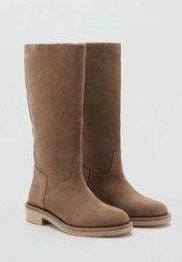 Botas altas de ante marrón hasta la rodilla con punta redonda y una suela de goma beige baja. Presentan un diseño simple con mínimos detalles de costura.