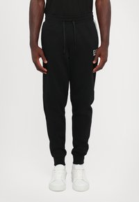 Svart joggebukser laget av myk stoff, med elastisk midje med snor, sidelommer og ribbestrikkede mansjetter. Hvite joggesko kompletterer stilen.