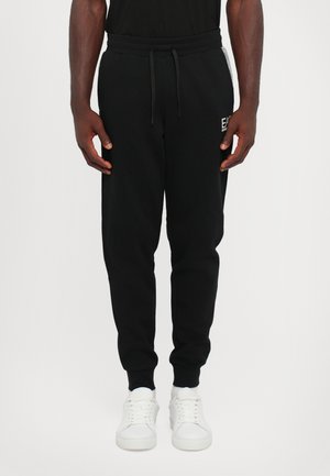 Black sweatpants made of soft fabric, featuring an elastic waistband with drawstring, side pockets, and ribbed cuffs. White sneakers complement the look.
