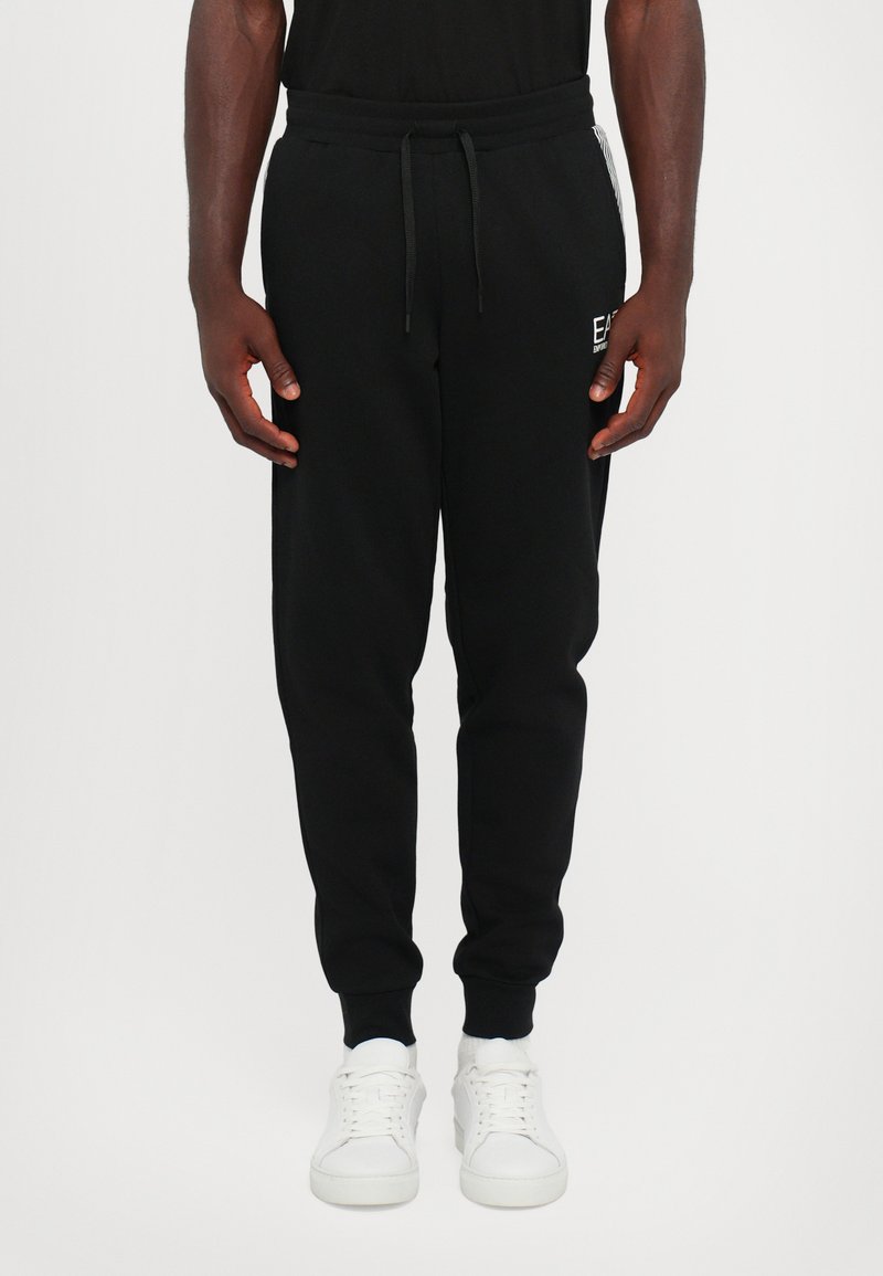 Svart joggebukser laget av myk stoff, med elastisk midje med snor, sidelommer og ribbestrikkede mansjetter. Hvite joggesko kompletterer stilen.