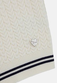 Suéter de punto de cable en color crema con una etiqueta en forma de corazón plateada y rayas marinas en el dobladillo. Textura suave con un diseño clásico.