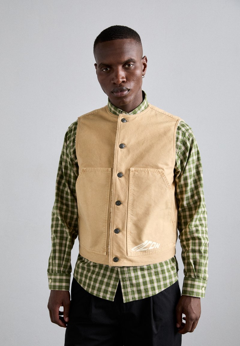 Tan canvas vest with a high collar, front buttons, and two large pockets, worn over a green and white checkered shirt.