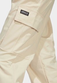 Pantalones cargo beige hechos de tejido ligero, con un gran bolsillo lateral y una sutil textura en forma de cuadrícula. Logotipo de Adidas Adventure visible.