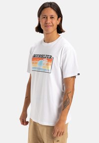 T-shirt bianco in cotone con un vivace design grafico che presenta onde, un sole e palme. Maniche corte e vestibilità rilassata.