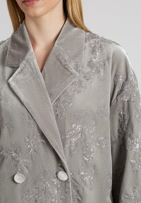 Blazer de terciopelo gris con bordados florales y lentejuelas, que presenta un cuello con muesca y dos botones circulares en el frente.
