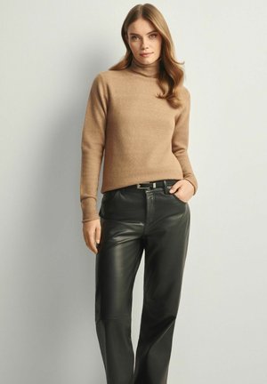 Karmelowy sweter z golfem z kaszmiru z prążkowanymi mankietami, zestawiony z gładkimi czarnymi skórzanymi spodniami. Modelka stoi na tle neutralnym.