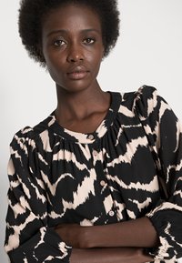 Blusa estampada em preto e bege com decote redondo, mangas bufantes e fecho frontal com botões. Tecido leve com acabamento texturizado.