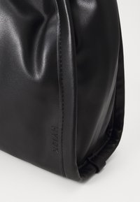 Zwarte faux lederen tas met een gladde oppervlakte, zachte plooien en een opvallend logo dat aan de zijkant is geperst. Compacte, rechthoekige vorm.