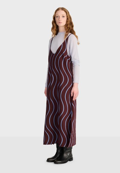 Femme aux longs cheveux bouclés portant un haut gris clair à manches longues sous une robe marron sans manches avec des rayures ondulées bleues et des bottines noires.