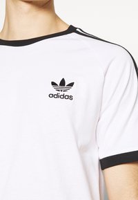 Bílý tričko s černým kontrastním límečkem a okraji rukávů. Na levé části hrudníku je černě vyšitý logo Adidas ve tvaru trojlístku.