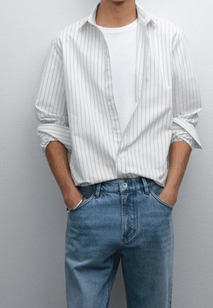 Mand iført en hvid t-shirt med en hvid, tyndstribet skjorte over og blå jeans, hænder afslappet i lommerne mod grå baggrund.