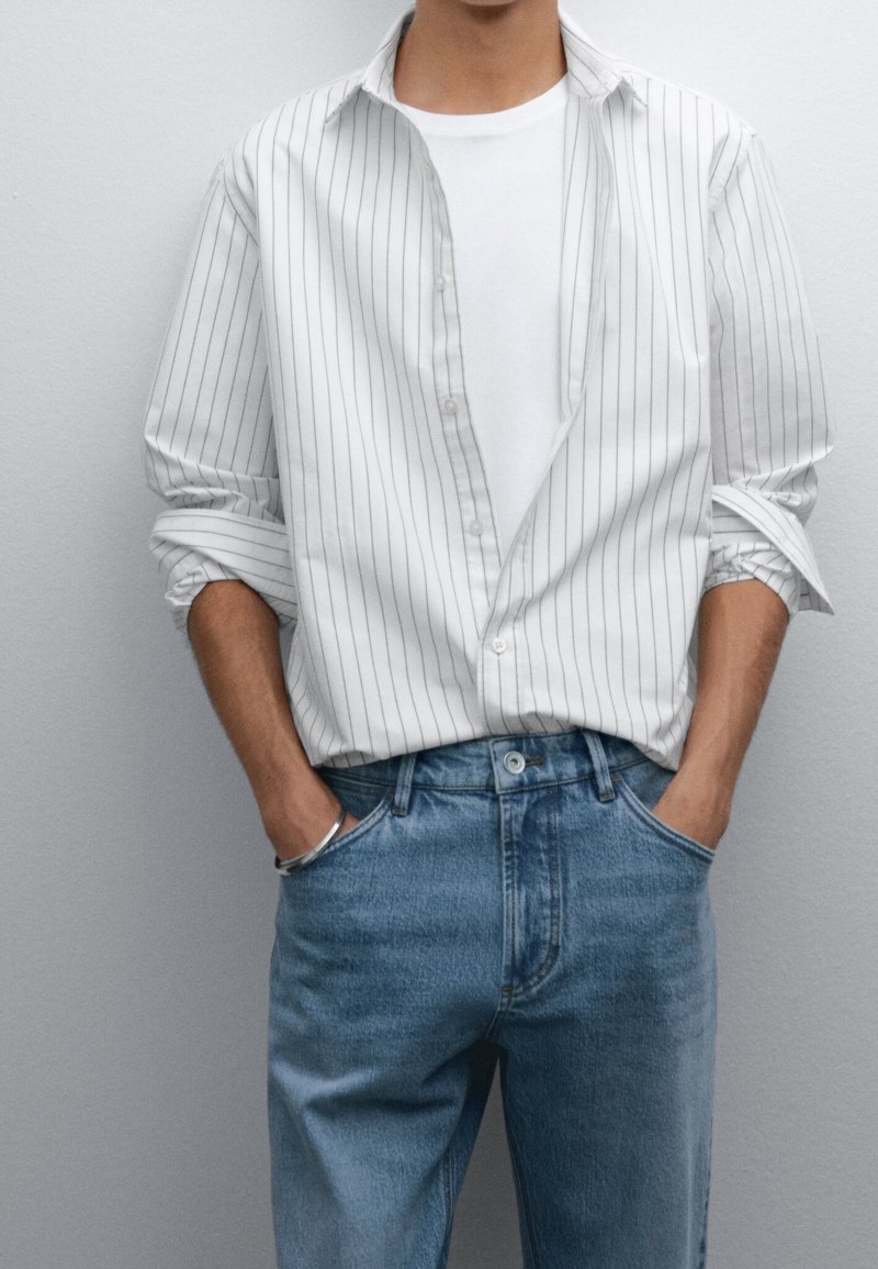 Homme portant un t-shirt blanc avec une chemise blanche à fines rayures par-dessus et un jean bleu, mains décontractées dans les poches sur fond gris.