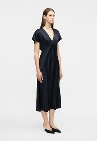 Γυναίκα στέκεται φορώντας μαύρο σατέν midi φόρεμα με κοντά μανίκια και βαθύ V λαιμόκοψη, συνδυασμένο με μαύρα μυτερά παπούτσια με χαμηλό τακούνι.