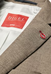 Brauner strukturierter Blazer mit einem roten Lippen-Revers-Anstecker und einem rot-weißen Etikett mit der Aufschrift "DIGEL The Menswear Concept Vintage."
