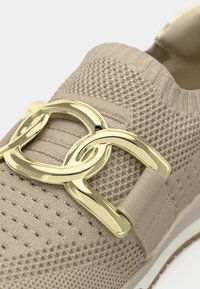 Μπεζ πλεκτό αθλητικό παπούτσι με μεγάλη χρυσή αλυσίδα, μαλακή υφασμάτινη επάνω επιφάνεια και ελαστική πλαϊνή επένδυση για εύκολη φόρμα.