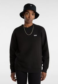 Zwarte sweatshirt met een ronde hals en geribbelde boorden, met een witte Vans-logo op de borst. Gecombineerd met een zwarte bucket hat.