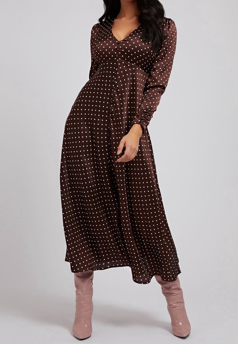 Femme portant une robe midi marron à manches longues avec des pois blancs, associée à des bottes à talons hauts rose clair, debout devant un fond gris.