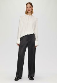 Weiße Bluse mit gerafftem Ausschnitt, langen Ärmeln und Schlitzdetail, kombiniert mit schwarzen, weiten Satin-Hosen und chunkigen schwarzen Schuhen.