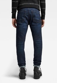 Jeans de mezclilla azul oscuro con un corte ajustado, que presentan costuras sutiles en los bolsillos traseros y dobladillos enrollados sobre botas negras.