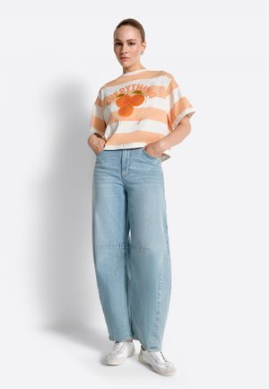 Moteris stovinti su rankomis kišenėse, dėvi persikinės spalvos ir balto dryžuoto marškinėlio su apelsino vaisiaus grafika ir plačias šviesiai mėlynas džinsines kelnes bei baltus sportinius batelius.