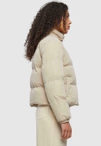 Veste rembourrée beige avec un col montant, design matelassé, poignets élastiques et poches latérales, montrée de côté sur un fond neutre.