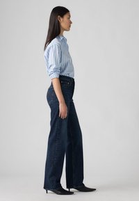 Sieviete svītrainā zilā krekla ar sarullētām piedurknēm, tumšās platās biksēs un melnās norādītajās papēžos stāv sāniski pret neitrālu fona.