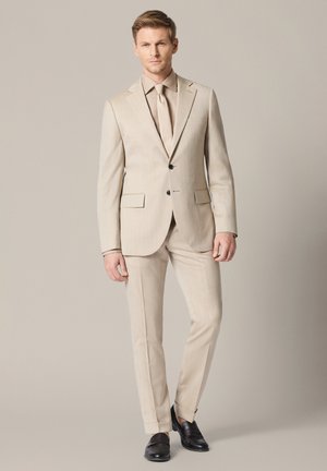 Mężczyzna stojący w beżowym garniturze z dopasowaną koszulą i krawatem, w czarnych eleganckich butach, na jednolitym beżowym tle.