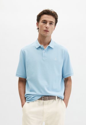 Młody mężczyzna o jasnobrązowych włosach, ubrany w jasnoniebieską koszulkę polo, beżowe spodnie i brązowy pasek, stojący z rękami w kieszeniach na białym tle.