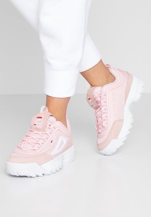 Osoba nosící bílé kalhoty a růžové robustní tenisky Fila s tlustými bílými podešvemi na světle šedé podlaze.