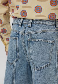 Jeans in denim blu chiaro con vita alta, dotati di due tasche posteriori, cuciture visibili e una superficie texturizzata. Camicia a motivi parzialmente visibile.