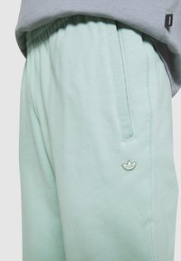 Ljust gröna träningsoverallsbyxor med elastisk midja, sidoficka och en liten vit broderad Adidas-logotyp nära fickan. Grå topp delvis synlig.