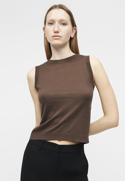 Νεαρή γυναίκα με ίσια μαλλιά, φοράει αμάνικο καφέ πλεκτό τοπ και μαύρο παντελόνι, στέκεται μπροστά σε απλό φωτεινό φόντο.